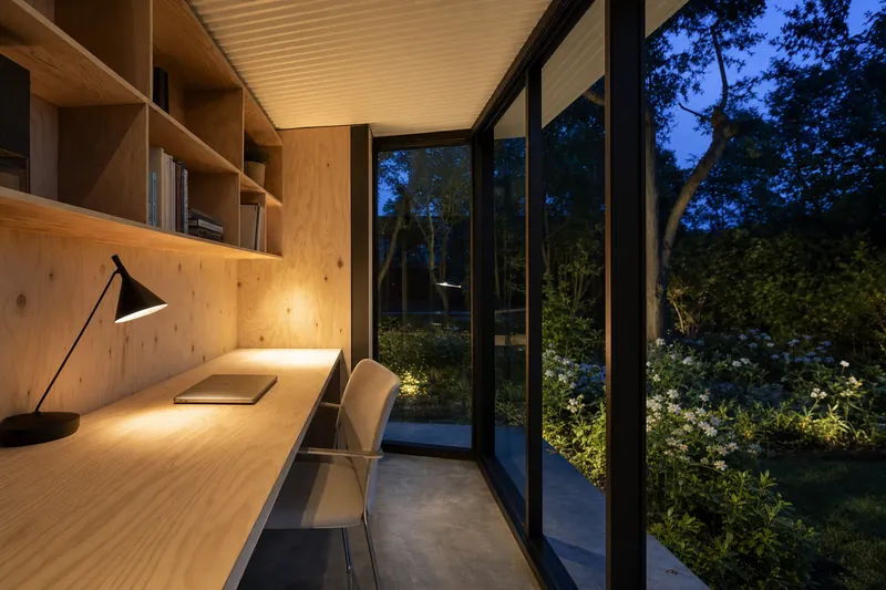 Escritório de jardim iluminado ao anoitecer, visto do interior para o jardim