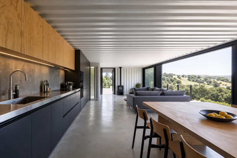 Container home interior with a dark galley kitchen, long timber table and living area beyond